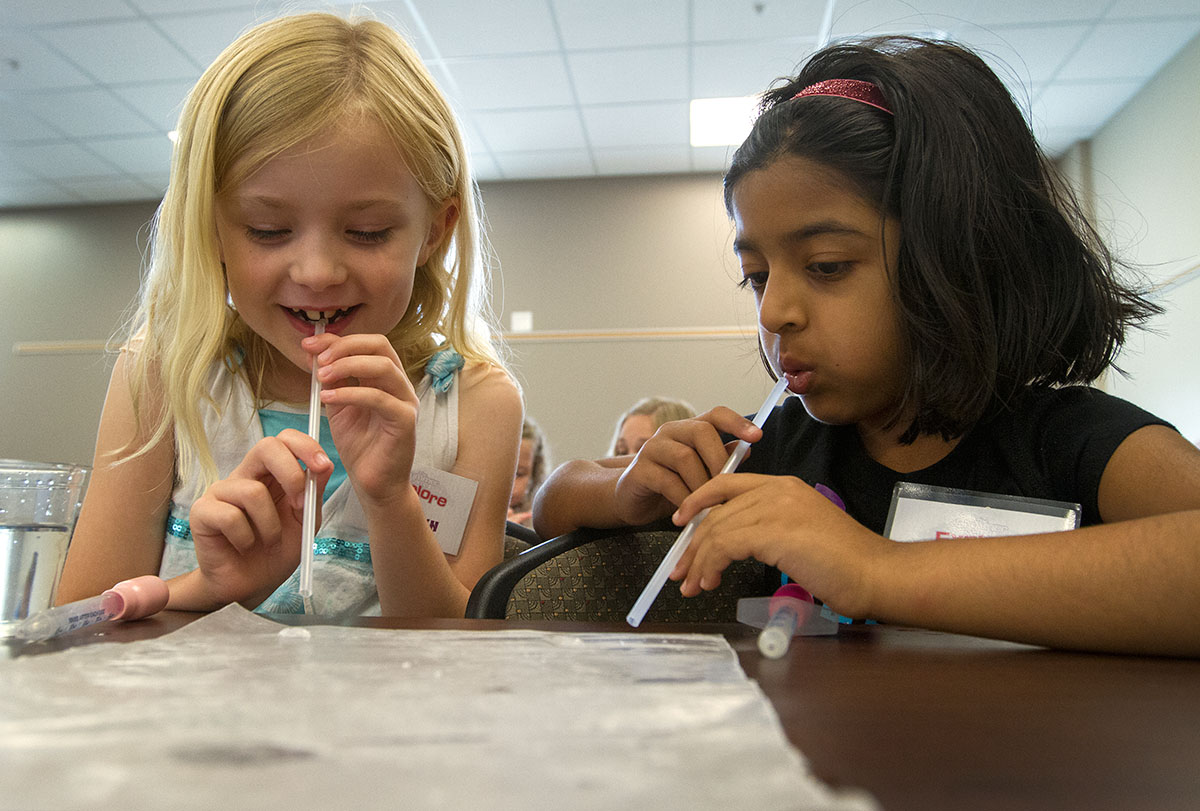Caitlyn (left) and Neha race to blow water droplets across wax paper in Science Monday, July 6, during Camp Explore. (Photo by Sam Oldenburg)