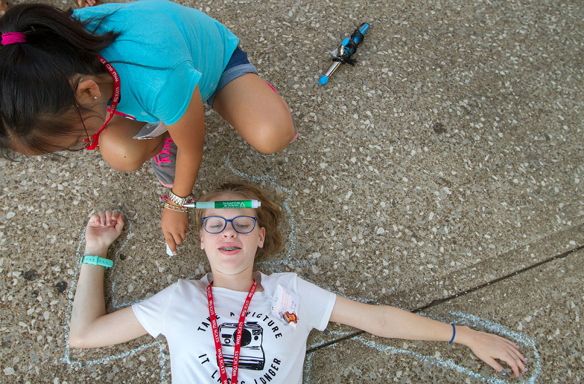 Lillie Green of Bowling Green draws a chalk outline around Grace Theilen of Hendersonville, Tenn., during the optional CSI: Bowling Green, Chalk Scene Investigations Monday June 19. Campers were given an assortment of props then had to come up with a crime story involving the items. (Photo by Sam Oldenburg)